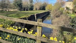 Un canal surmonté d’un pont de pierre, une clôture en bois et, au premier plan, des jonquilles jaunes.