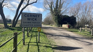 Un panneau indiquant l’entrée de Cerney Wick, un village de la paroisse saxonne, mentionnant la charte de l’an 999, avec une route de campagne et des arbres en arrière-plan.