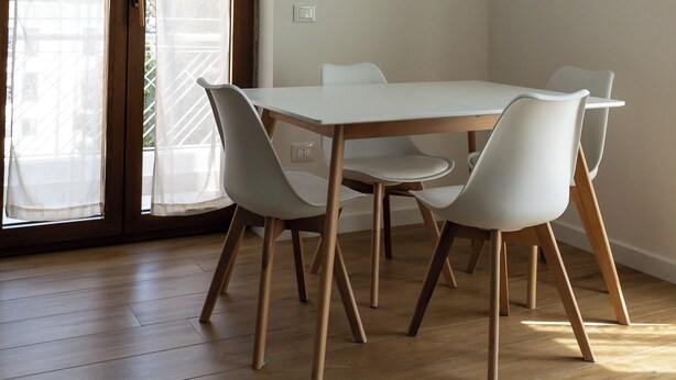 Un coin repas avec une table blanche et quatre chaises, un plancher en bois et une porte-fenêtre coulissante donnant sur un balcon extérieur.