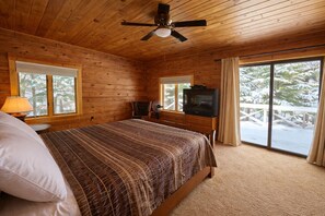 Une chambre dans une cabane en bois, avec un lit, un ventilateur de plafond, une télévision et de grandes fenêtres donnant sur un paysage enneigé.