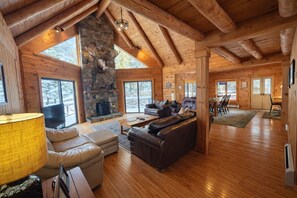 A log cabin interior with a stone fireplace, leather furniture, and large windows. - Waterfront Cabin perfect for Families, full lake access (Northwest Piscataquis)