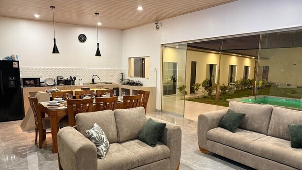 A modern living room with a dining area, a kitchen, and a glass wall leading to a pool. - Casa en Ica con piscina, parrilla y gran jardín (Ica)