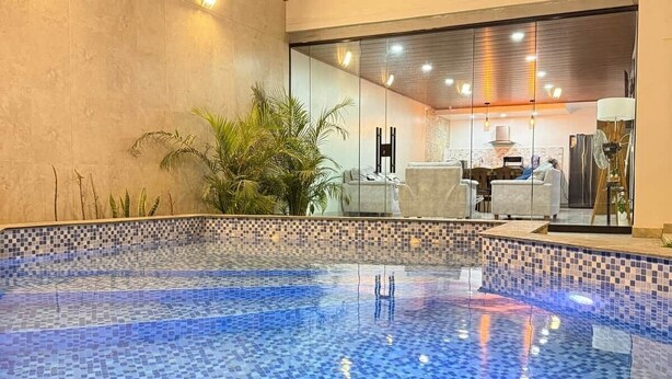 A swimming pool with mosaic tiles, a seating area with a table and chairs, and a view of a room with a large window.