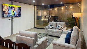 A modern living room with a flat-screen TV, grey sofas, a glass coffee table, and a view of the outdoors through large glass doors.