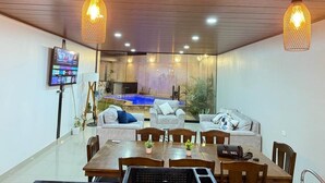 A modern living room with a dining area, a TV, and a pool in the background.