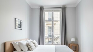Quarto moderno com uma cama grande, uma pequena mesa lateral, mesa de cabeceira e janela com vista para prédios.