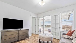 A modern living room with a flat-screen TV, a grey sofa, a round ottoman, and a wooden floor. - Big Foot Ranch Retreat – Spacious Idaho Home w/ View (Melba)
