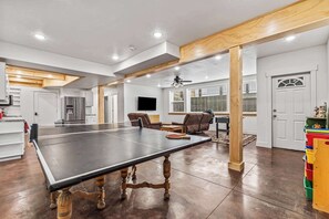 A spacious living area with a ping pong table, wooden beams, a ceiling fan, and a kitchen in the background.