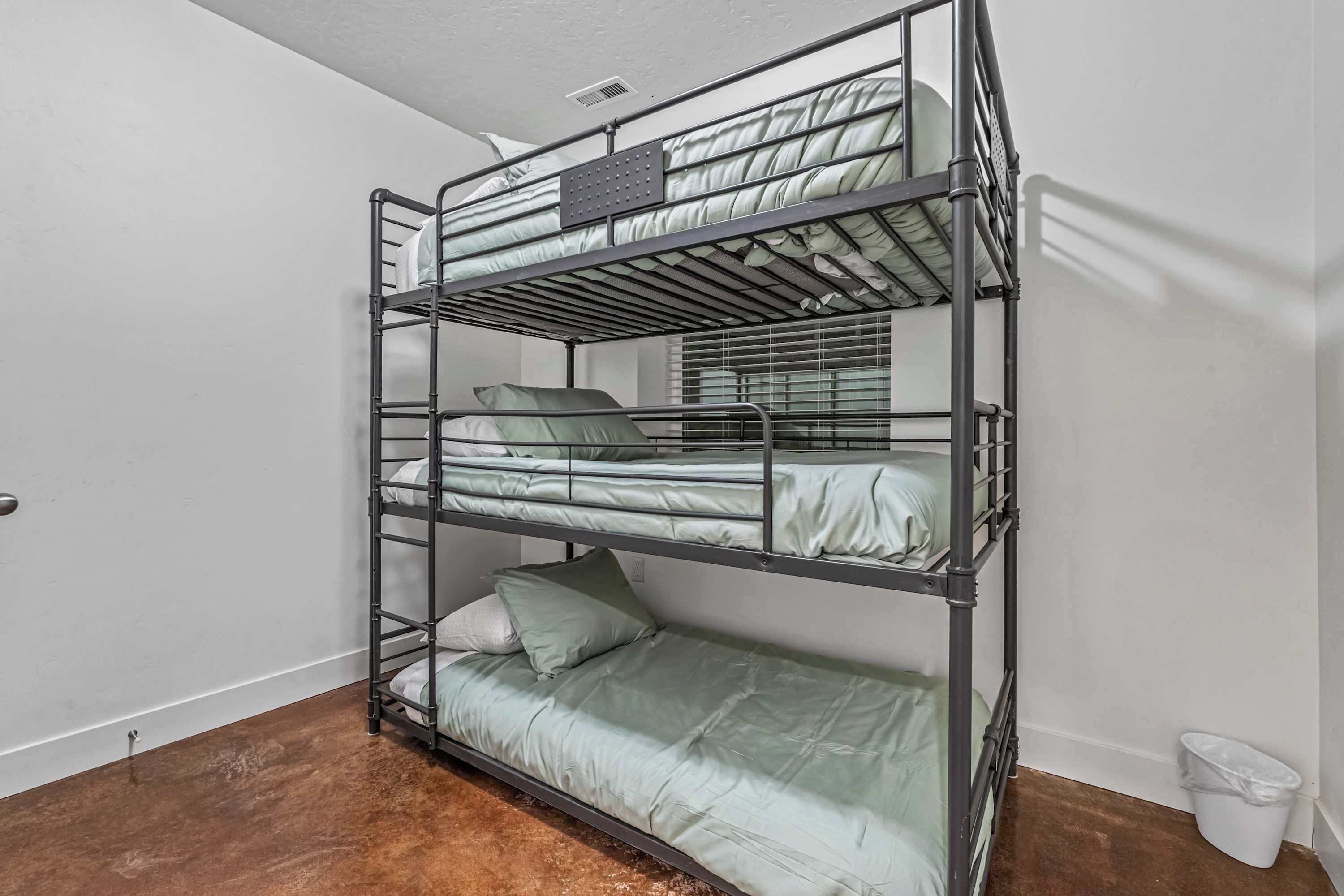 A bunk bed with three levels, a white wall, a window with blinds, and a white trash bin.