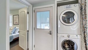 Un lavadero con lavadora y secadora de carga frontal, una puerta con ventana, y un dormitorio con cama y un cuadro enmarcado.