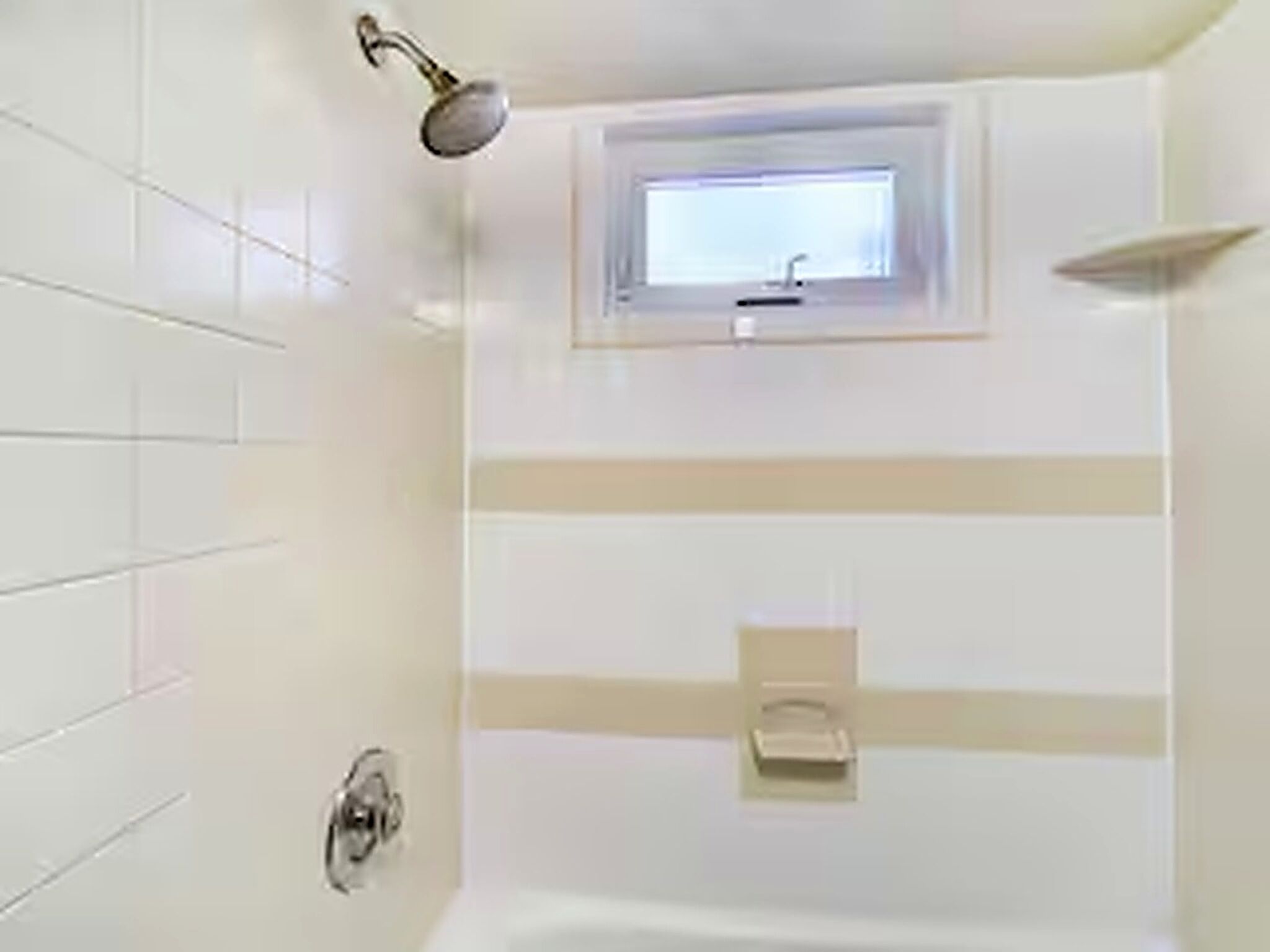 Baño con azulejos blancos, ducha, ventana y toallero.