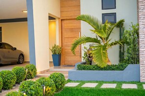 A modern building with a car parked in the driveway, a wooden door, and a well-maintained garden with various plants. - Villa Shorys: Free Electricity | 12Pax | Pool+Jacuzzi | A/C | Covered Parking (Punta Cana)