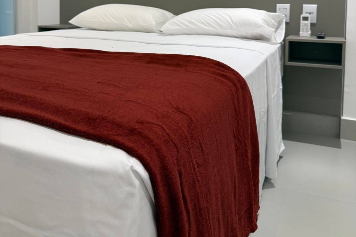 Une chambre d’hôtel avec un lit, du linge de lit blanc, une couverture rouge, une tête de lit grise et une table de chevet avec une lampe.