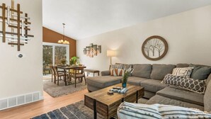 Ein Wohnzimmer mit einer Eckcouch, einem Couchtisch, einem Essbereich mit Tisch und Stühlen sowie einem großen Fenster.