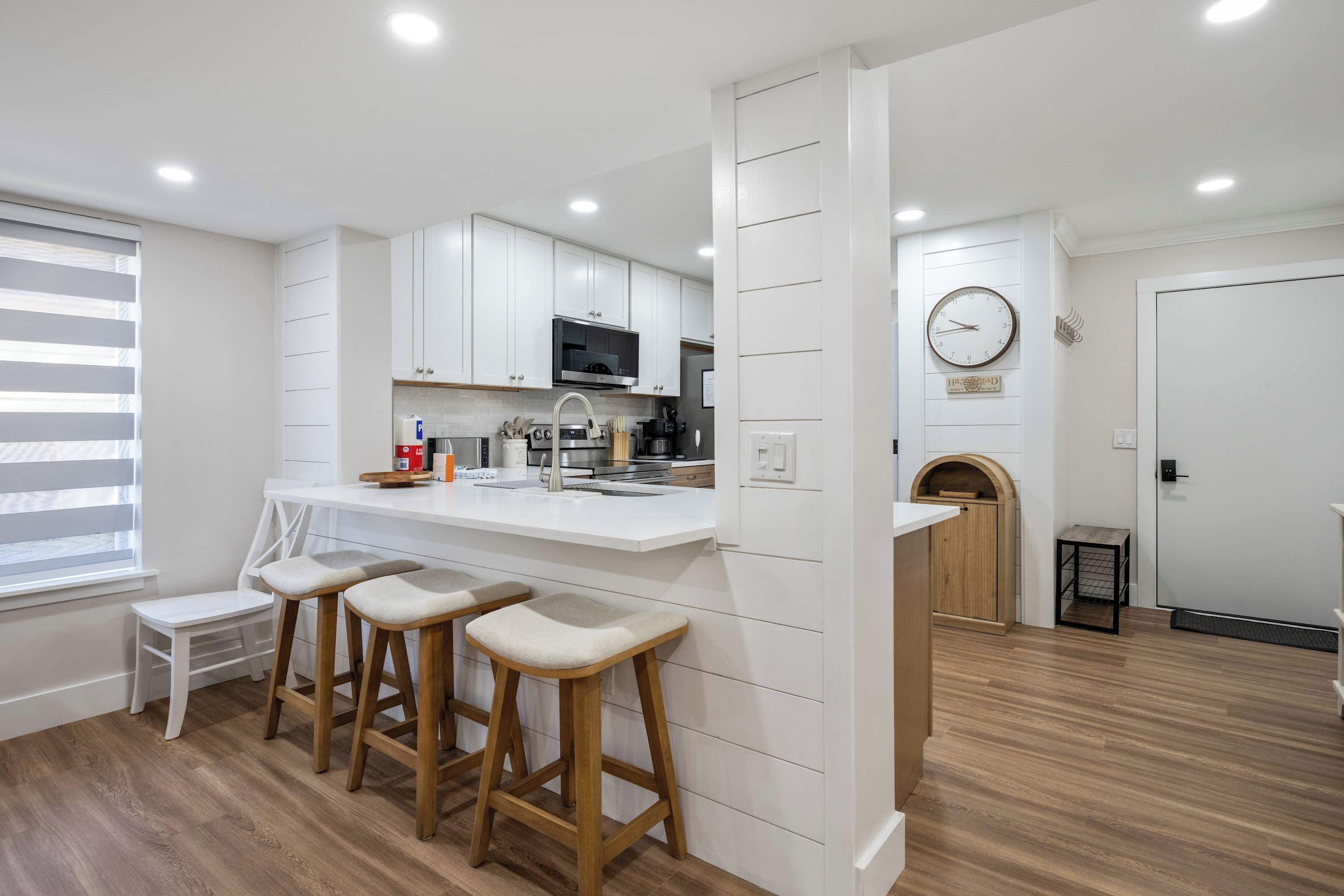 Une cuisine moderne avec des meubles blancs, un îlot central doté de tabourets, un four à micro-ondes et une horloge murale.