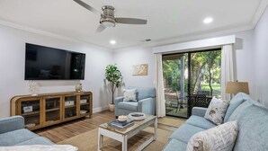 Una sala de estar con una televisión de pantalla plana, ventilador de techo, sofá, mesa de centro y puerta corrediza de vidrio que da a un área exterior.