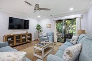A living room with a flat-screen TV, a ceiling fan, a sofa, a coffee table, and a sliding glass door leading to an outdoor area. - Courtside 1 Newly Renovated 2 BR First Floor Villa Walk to Beach & Free Tennis (Hilton Head Island)