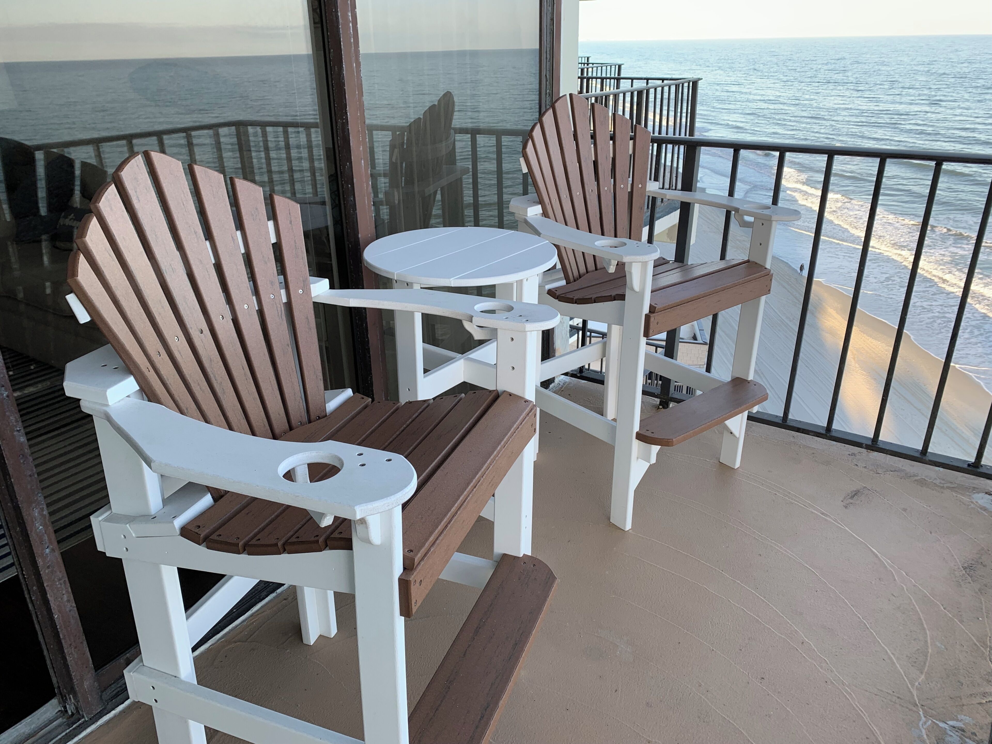 En balkon med to Adirondack-stole og et lille rundt bord, med udsigt over en strand.