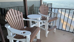 A balcony with two Adirondack chairs and a small round table, overlooking a beach.