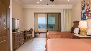 A hotel room with a bed, a desk with a television, a chair, a nightstand with a lamp, and a view of the sea through a sliding door. - Cozy 2 Bedroom Elite Suite at Villa del Palmar Loreto (Ensenada Blanca)