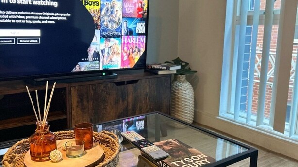 Una sala de estar moderna con un televisor de pantalla plana grande mostrando la interfaz de un servicio de streaming, una mesa de centro de vidrio con un control remoto, un libro y objetos decorativos.