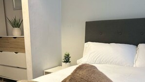 Una cama bien tendida con sábanas blancas y una toalla enrollada, una pequeña planta en la mesita de noche y un cabecero oscuro.