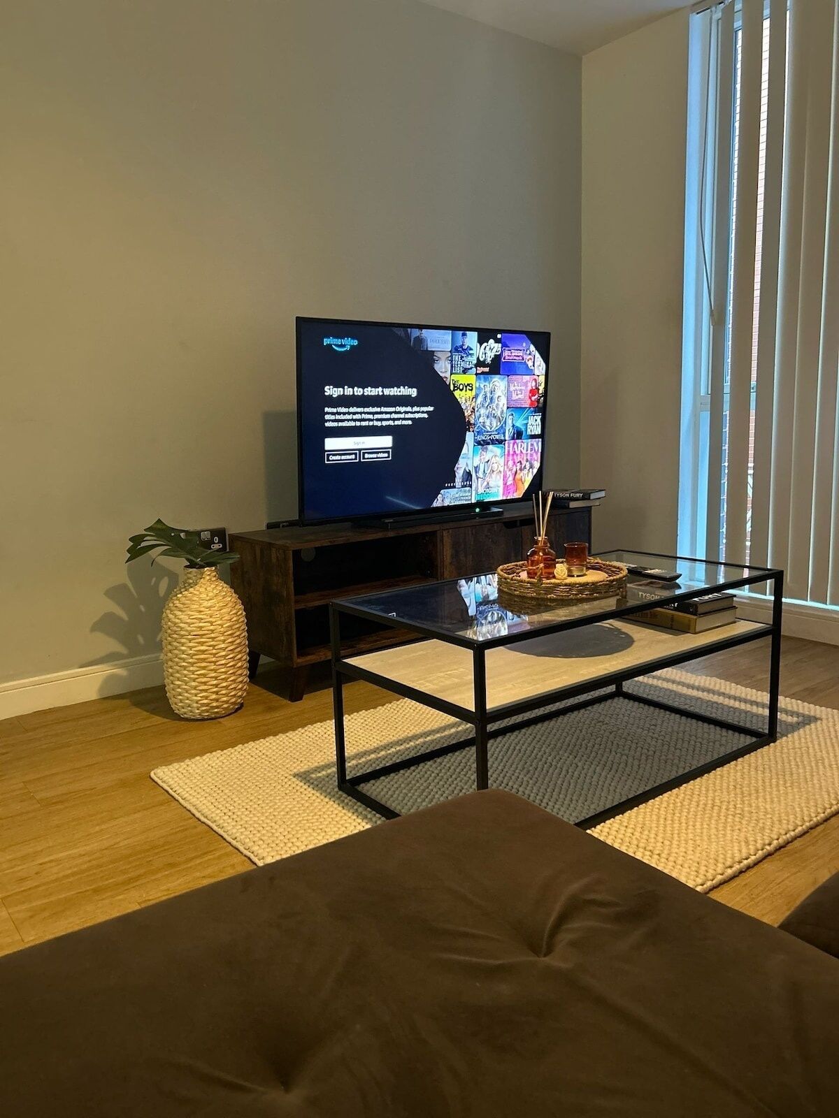 A living room with a television displaying a sign-in screen, a glass coffee table, a dark sofa, a woven vase, and a large window with blinds.
