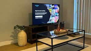 Un salón con una televisión mostrando una pantalla de inicio, una mesa de centro de vidrio, un sofá oscuro, un jarrón tejido y un ventanal con persianas.