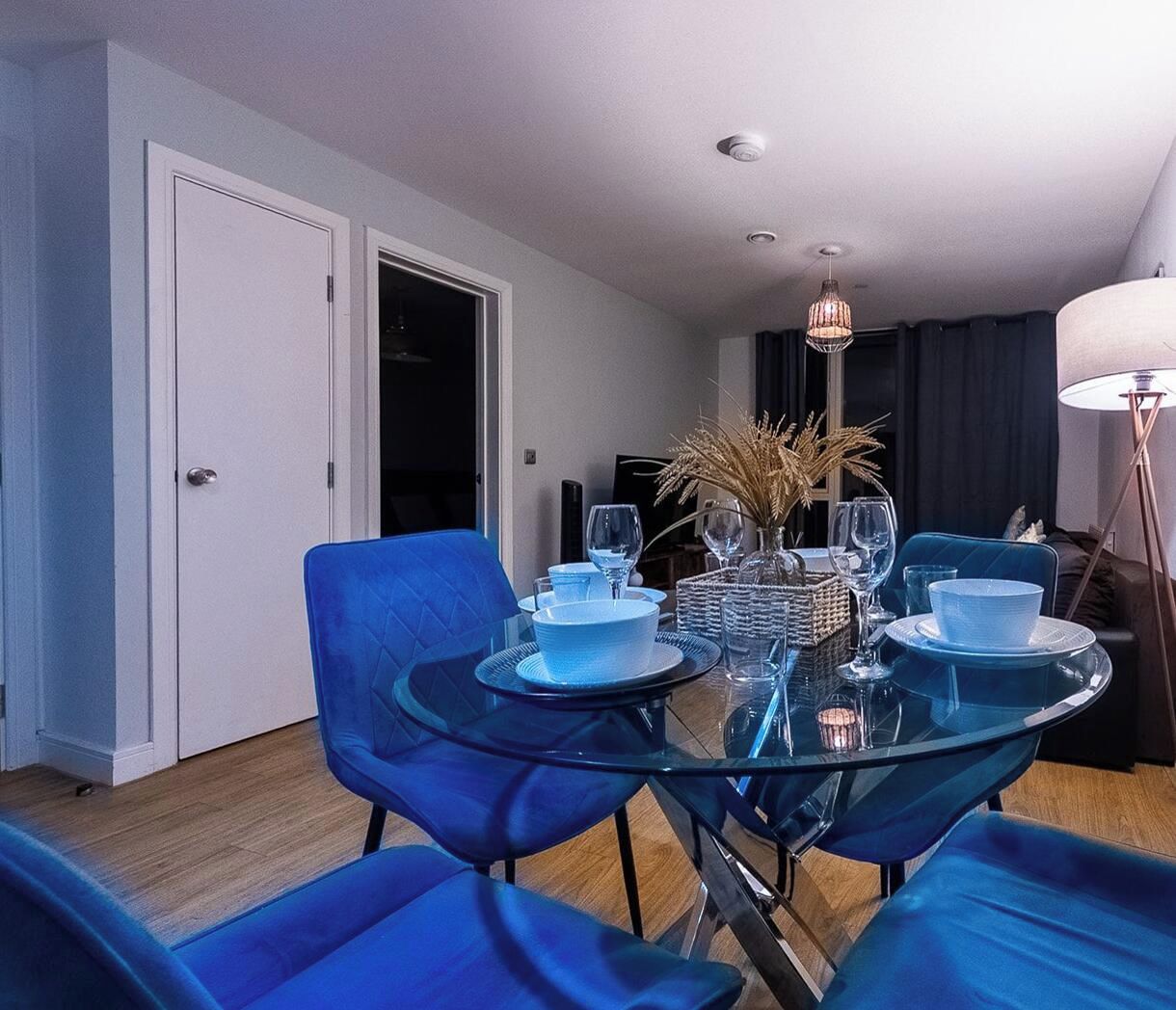 Un espace repas moderne avec une table en verre et des chaises bleues, une petite table d’appoint avec un vase, et une porte donnant sur une autre pièce.
