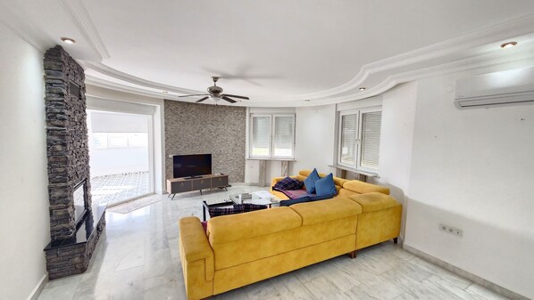 A living room with a yellow sofa, a stone fireplace, a flat-screen TV, and a ceiling fan.