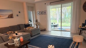 A living room with a sofa, a coffee table, a patterned armchair, and a view through a glass door to an outdoor area. - Beautiful 4-bedroom house with AC in Ormond Beach (Ormond Beach)