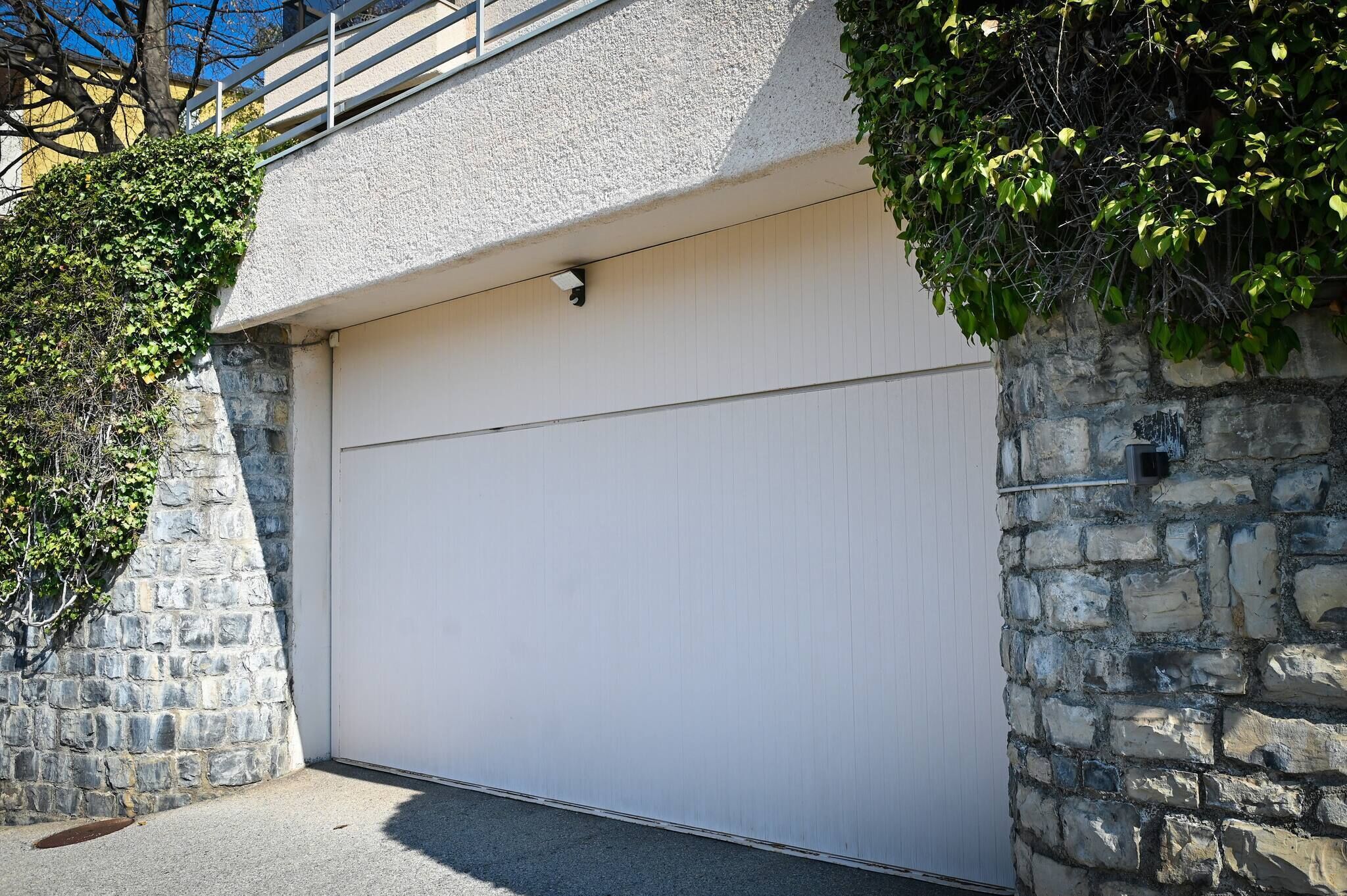 Un garage avec une porte blanche, situé à côté d’un mur en pierre et d’un arbre.