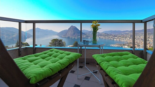 A balcony with two green cushioned chairs, a small table with a vase of flowers, and a scenic view of a lake and mountains. - Serenity Lakefront 2 Bedrooms Retreat in Lugano (Lugano)