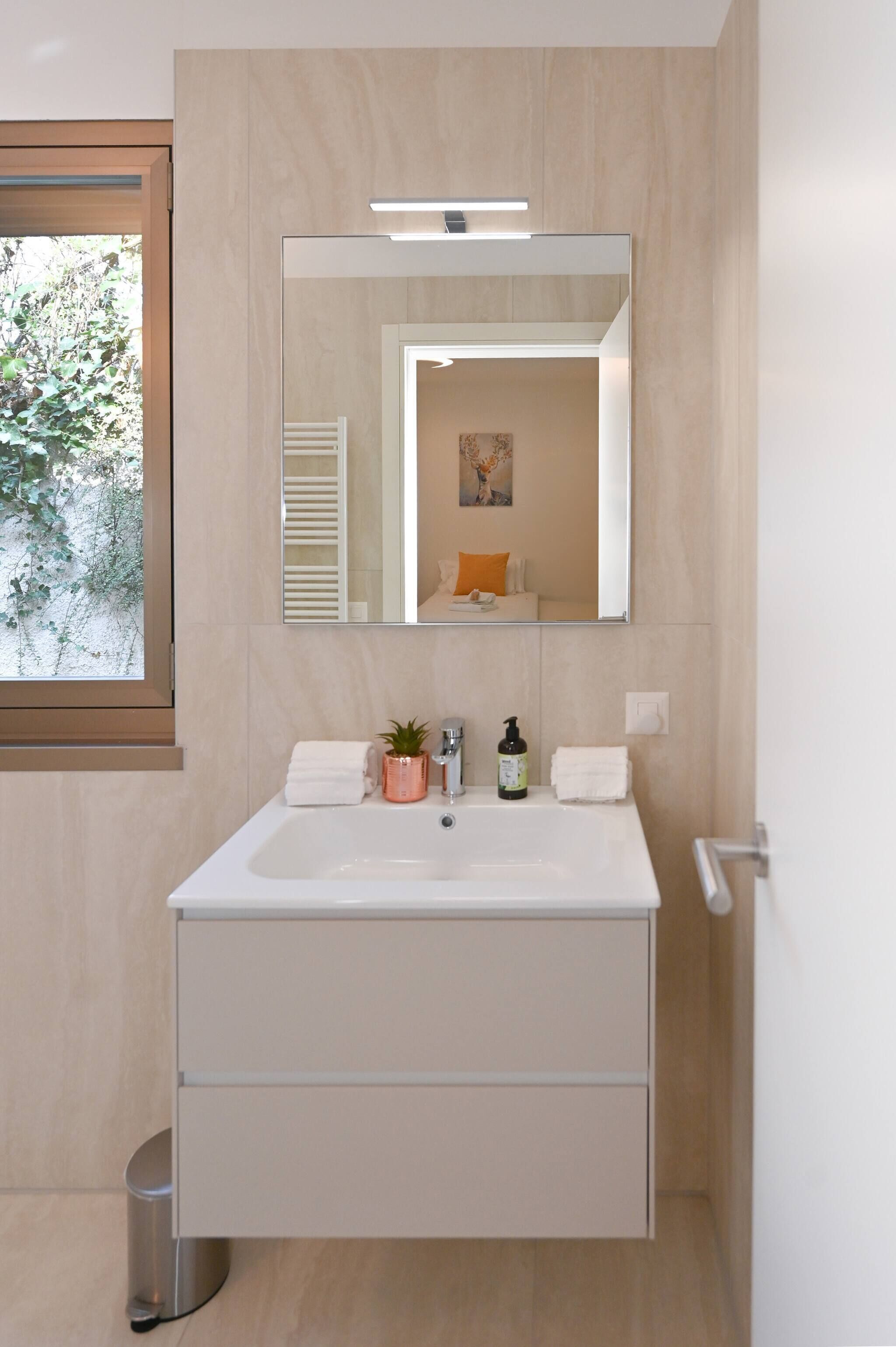 Ein modernes Badezimmer mit großem Spiegel, Waschbecken, Mülleimer und einem Fenster mit Blick auf Grünflächen.