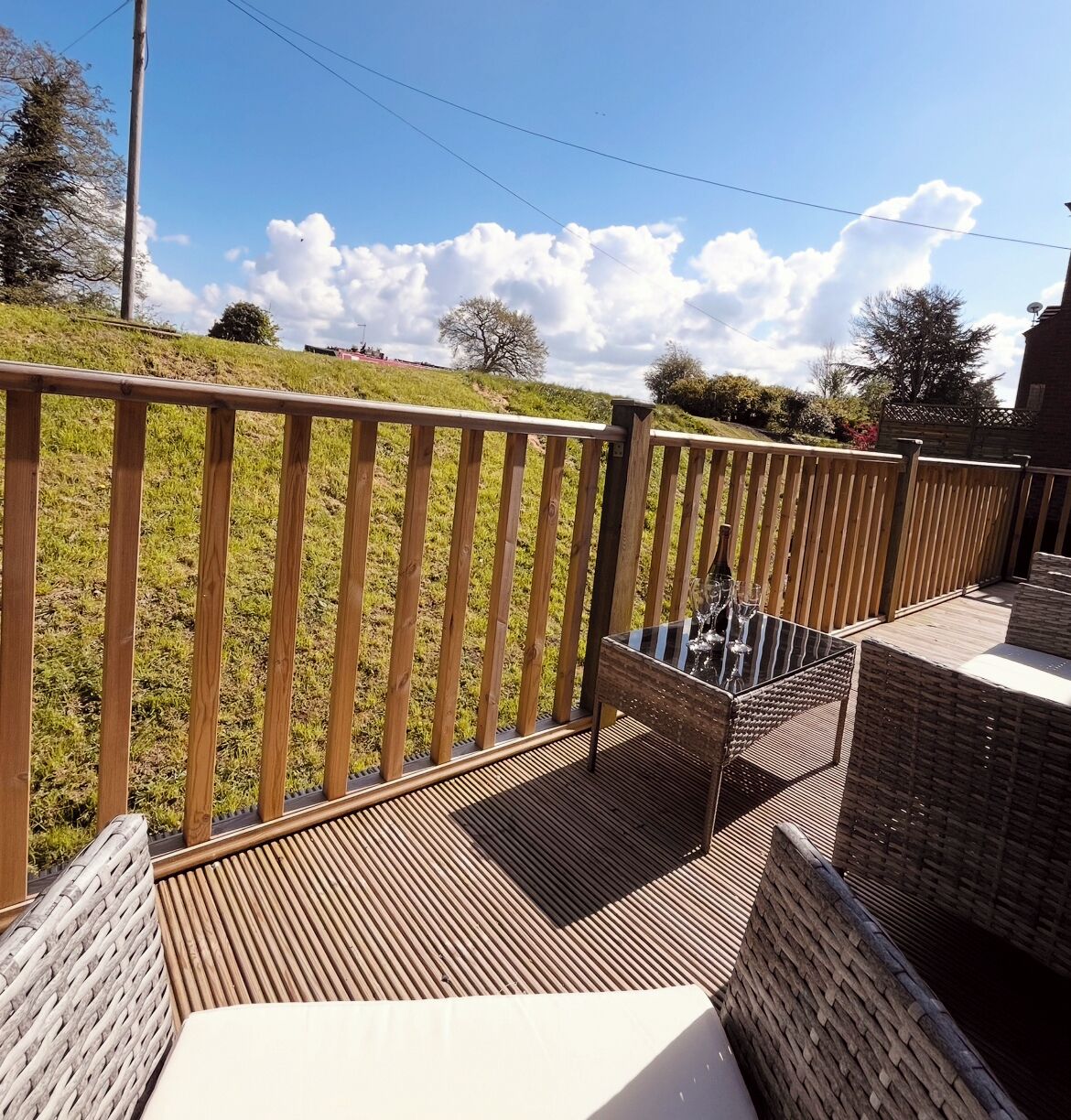 Una terraza de madera con mobiliario de mimbre y una mesa de cristal, con vistas a una zona verde con árboles.
