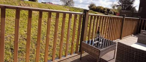 Una terraza de madera con mobiliario de mimbre y una mesa de cristal, con vistas a una zona verde con árboles.