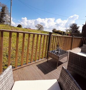 Una terraza de madera con mobiliario de mimbre y una mesa de cristal, con vistas a una zona verde con árboles.