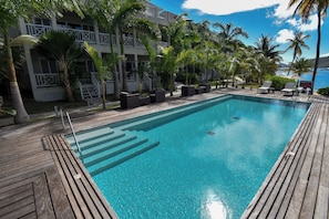 A rectangular swimming pool with clear blue water, surrounded by a wooden deck, palm trees, and a building with balconies. - Condo Style Waterfront Suites overlooking Falmouth Harbour marina (English Harbour Town)