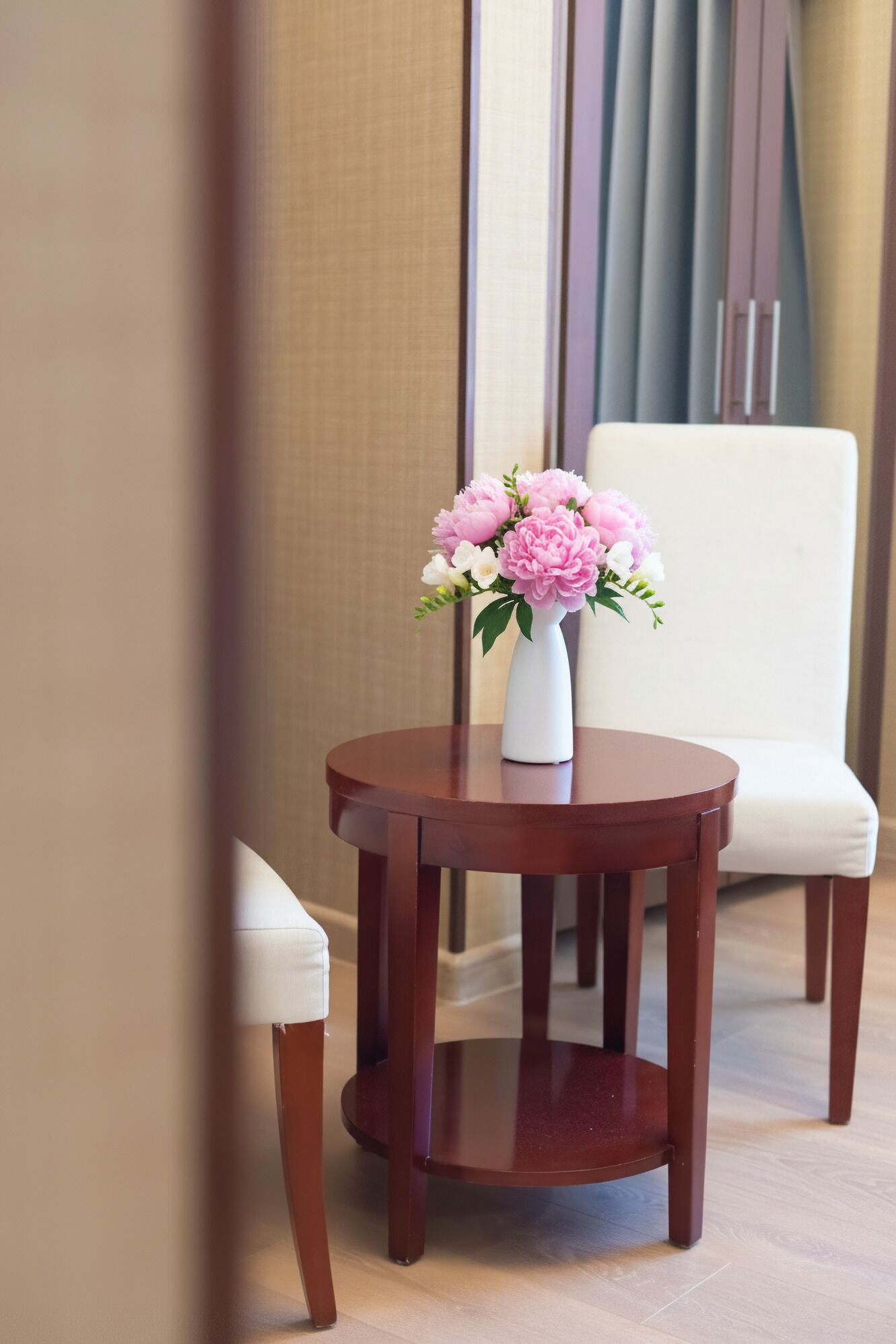Una pequeña mesa de madera con un jarrón blanco con flores rosadas, ubicada frente a una silla blanca en una habitación con alfombra color crema y un amplio ventanal con cortinas.