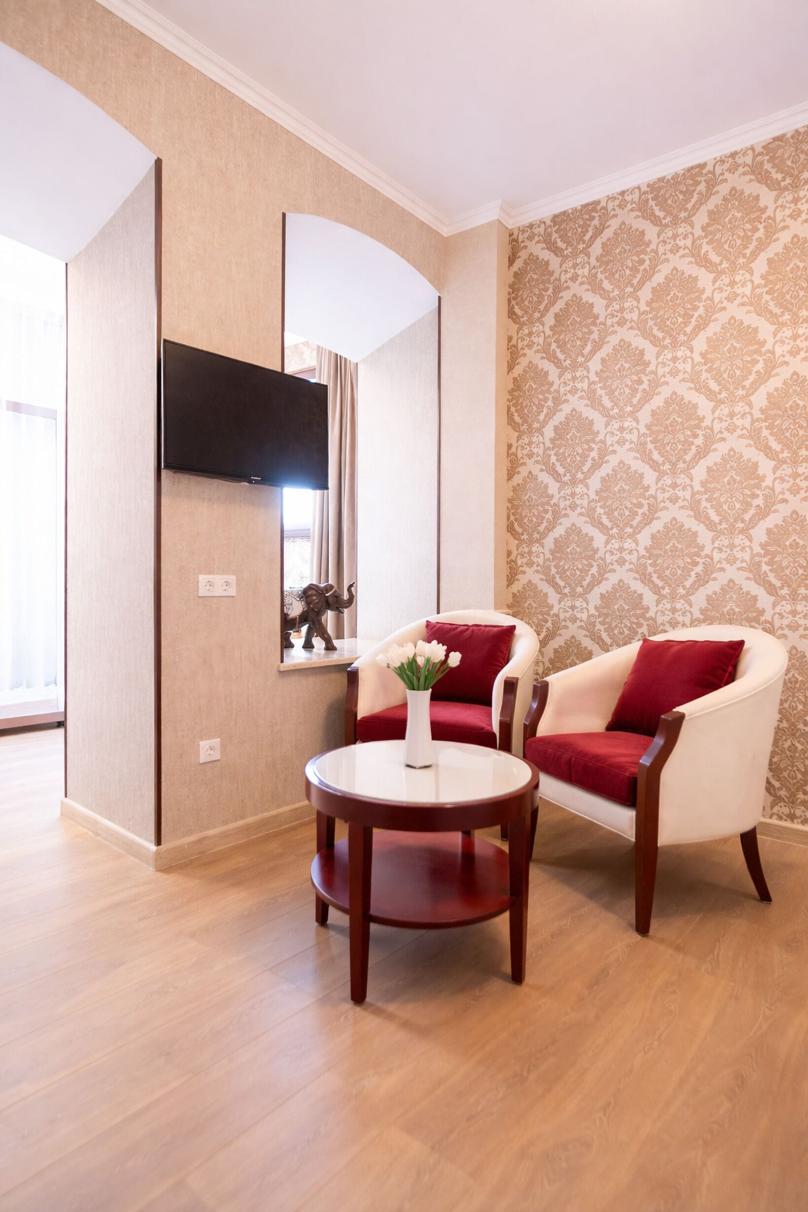 A room with a sofa, a small table with a vase, a flat-screen TV mounted on the wall, and patterned wallpaper.