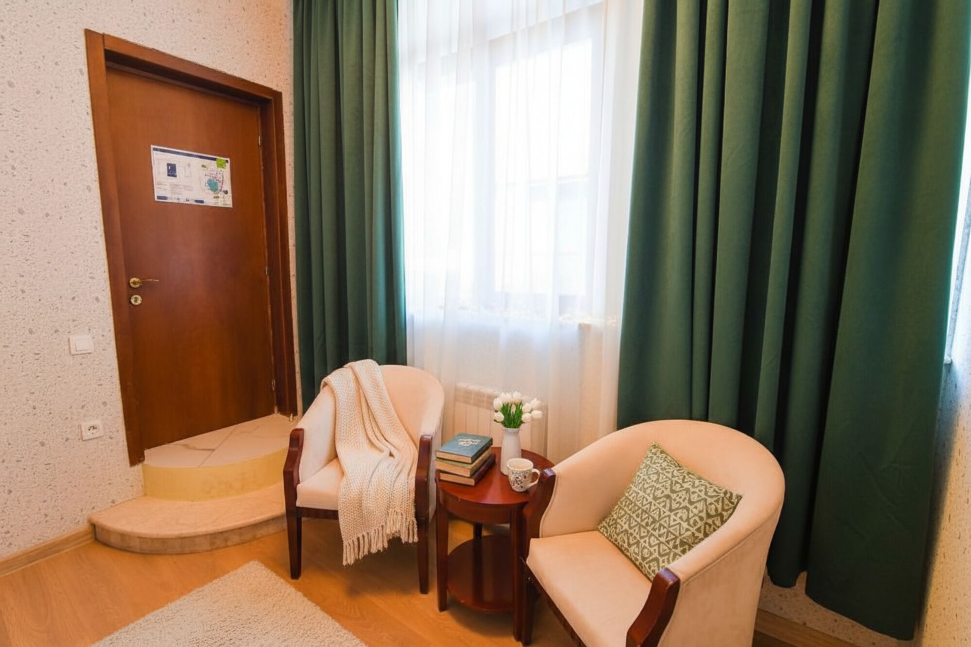 A room with a wooden door, a small table with a book and a mug, a beige armchair with a patterned pillow, and a window with green curtains.