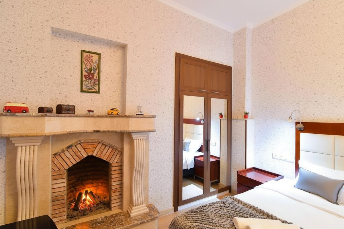 A bedroom with a fireplace, a bed, a nightstand, a mirror, and a door leading to another room.