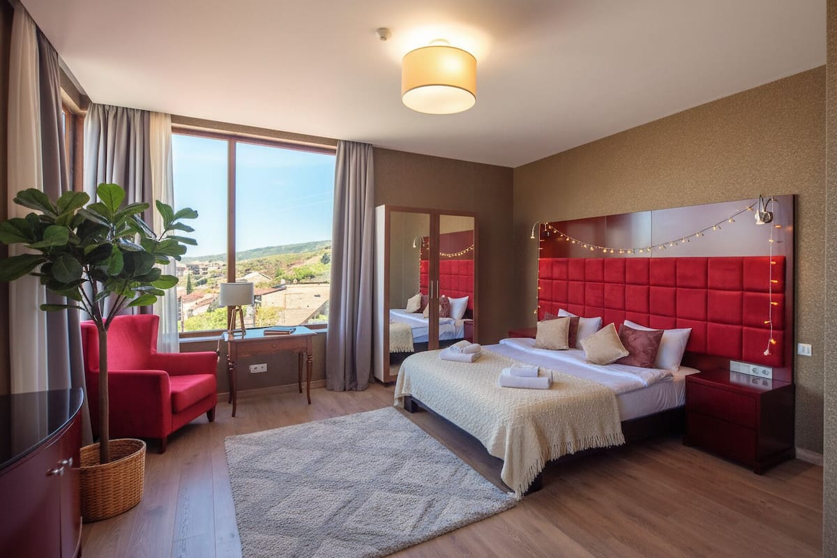 A hotel room with a large bed, a red armchair, a desk, and a view of the landscape.