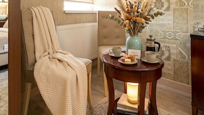 A cozy room with a chair, a table with a vase of flowers, a coffee maker, and books.