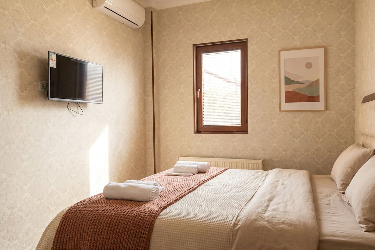 A hotel room with a bed, a flat-screen TV, a window with blinds, and a framed picture on the wall.