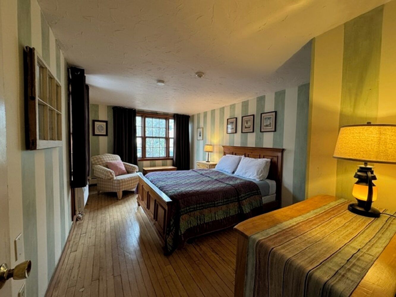 A hotel room with a wooden floor, a bed with a patterned bedspread, a chair, a nightstand with a lamp, and a window with curtains.