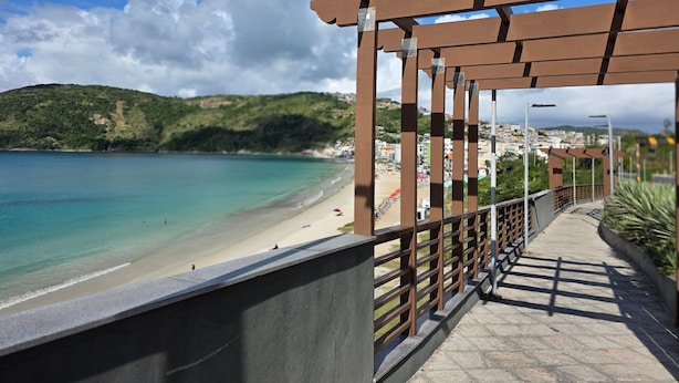 A coastal walkway with a view of a beach and hills. - Apartamento em Cond. de Luxo  (Arraial do Cabo)