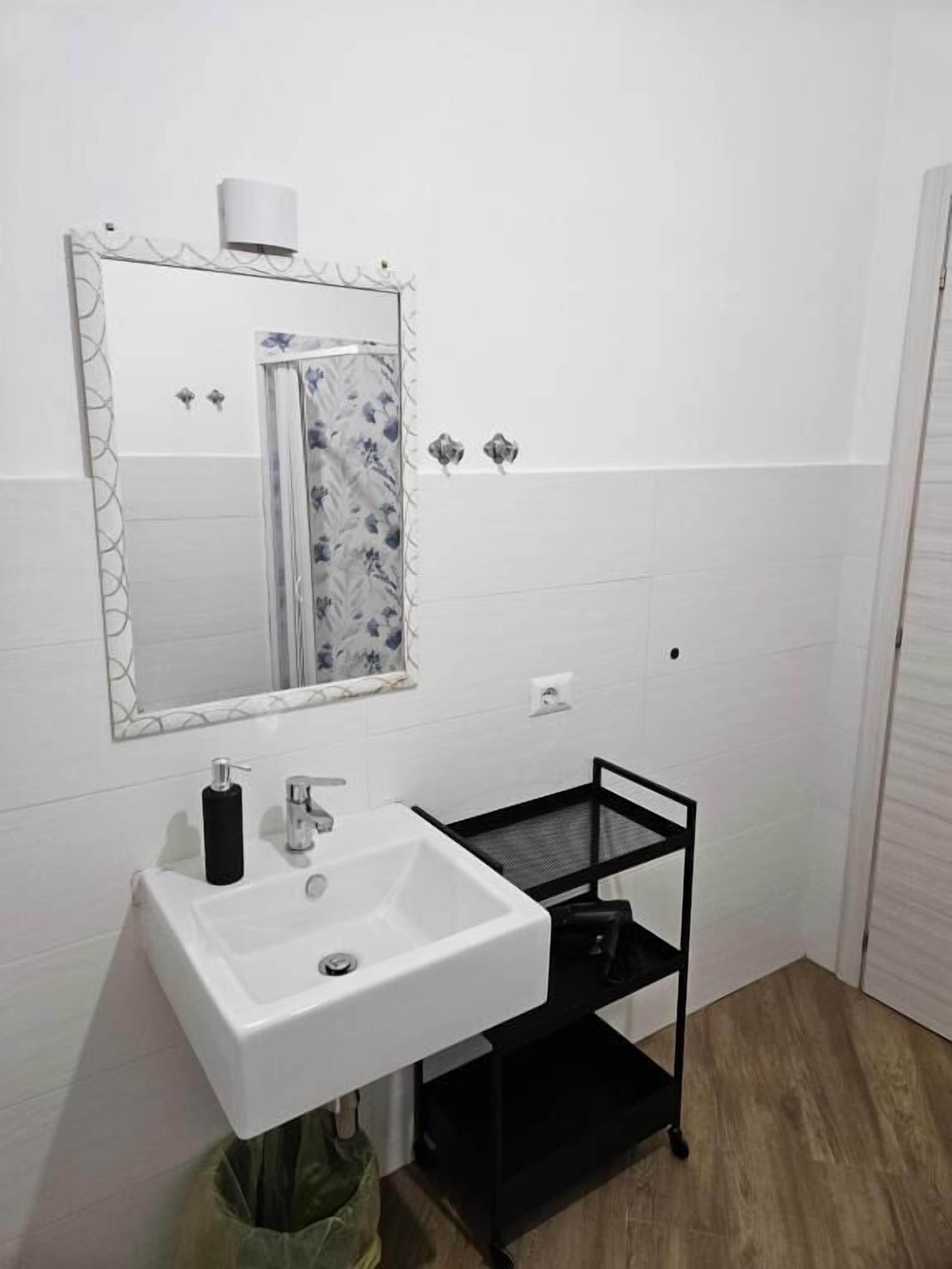 A bathroom with a white sink, a mirror, a black shelf, and a green trash bag.