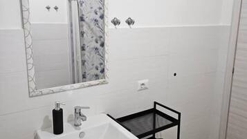 A bathroom with a white sink, a mirror, a black shelf, and a green trash bag.