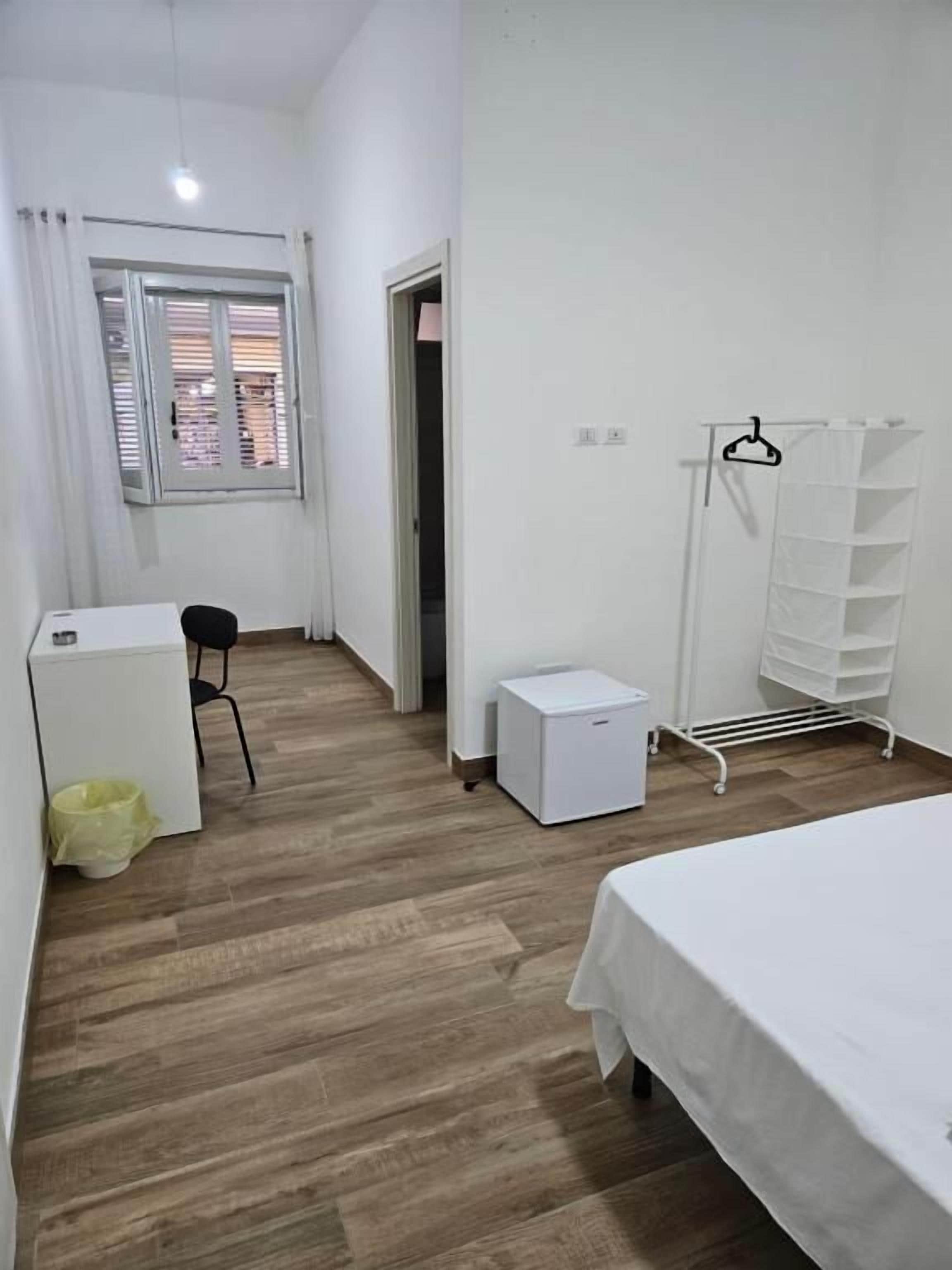 Een slaapkamer met een bed, een bureau, een stoel, een klein wit kastje, een droogrek en een raam met zonwering.